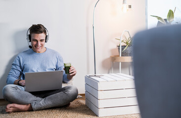 Smiling young man wearing wireless headphones watching video on laptop while sitting against wall in living room