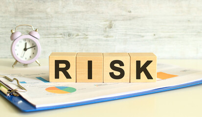 Wooden cubes lie on a folder with financial charts on a gray background. The cubes make up the word RISK.