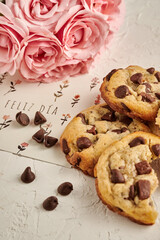 galletas de chocolate con chips  en una mesa blanca rustica con una tarjeta que dice feliz dia y rosas rosadas