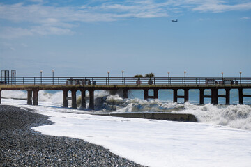 Fototapeta premium A long pier, under which big waves roll and an airplane in the sky