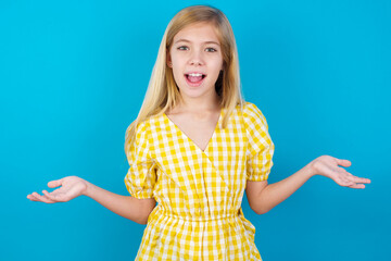 So what? Portrait of arrogant beautiful Caucasian little girl wearing yellow dress over blue background shrugging hands sideways smiling gasping indifferent, telling something obvious.