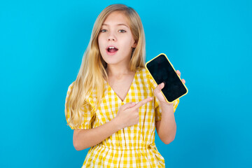 Excited beautiful Caucasian little girl wearing yellow dress over blue background holding and pointing with finger at smartphone with blank screen. Advertisement concept.