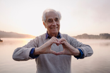 Smiling man making heart shape with hands while standing against lake