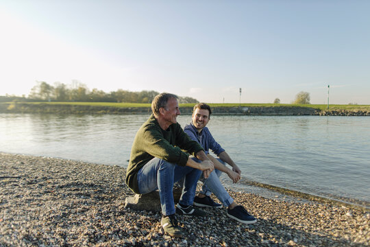 Father and son spending leisure time sitting at riverbank against clear sky