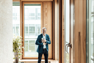 Smiling businessman with digital tablet standing against glass window in office corridor