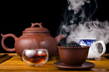 Chinese tea ceremony. Clay teapot with red tea Lapsang souchong on a black background, ceramic cup and jug of hot tea with vapour.