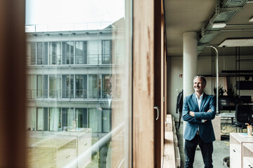 Contemplating businessman with arms crossed looking through window while standing in office