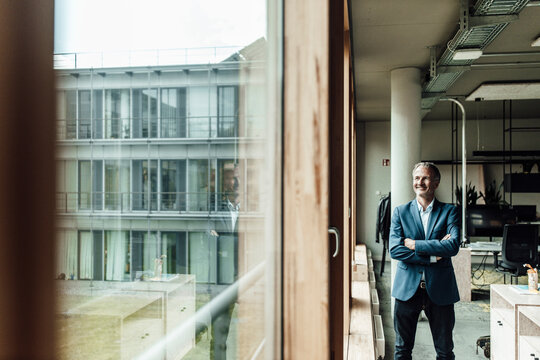 Contemplating businessman with arms crossed looking through window while standing in office