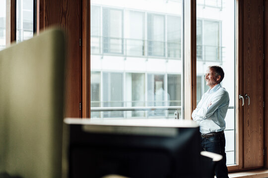 Smiling senior male entrepreneur with arms crossed looking through glass window in office