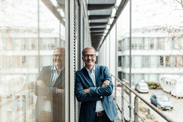 Smiling male entrepreneur with arms crossed leaning on window in office balcony