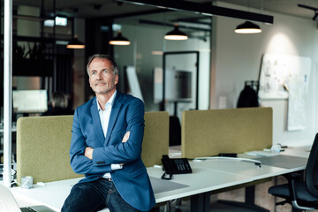Male business professional with arms crossed sitting on desk in office