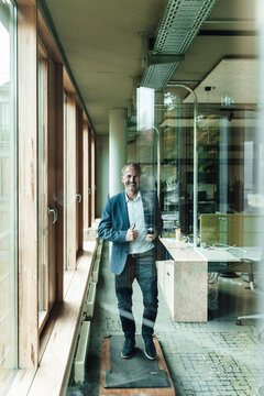 Smiling male senior entrepreneur exercising on treadmill seen trough glass window in office