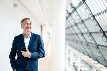 Smiling businessman with smart phone looking away while standing in office corridor