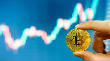 Investor hand holding gold Bitcoin with blurred candlestick chart in the background. BTC known as digital gold asset in cryptocurrency world.