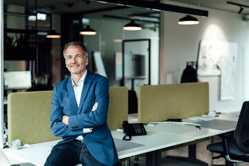 Smiling businessman with arms crossed sitting on desk in office