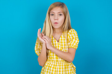 Surprised emotional beautiful Caucasian little girl wearing yellow dress over blue background rubs palms and stares at camera with disbelief