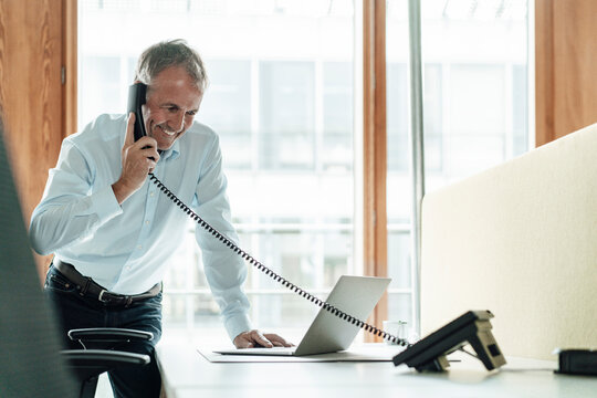 Smiling male entrepreneur using laptop while talking on telephone at desk