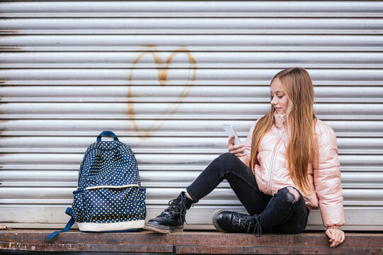 Girl with long brown hair looking at mobile phone while sitting against shutter