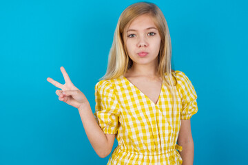 beautiful Caucasian little girl wearing yellow dress over blue background makes peace gesture keeps lips folded shows v sign. Body language concept
