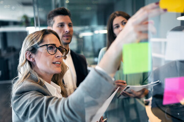 Businesswoman reading adhesive note on glass wall while standing with colleague at office