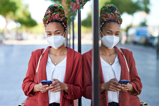 Woman wearing headscarf and protective face mask using mobile phone while leaning on glass wall