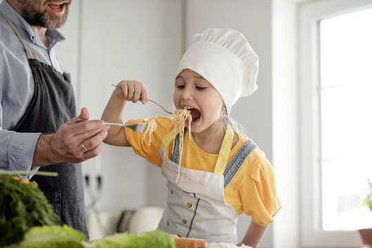 Girl wearing apron and chef's hat eating noodles with father while standing in kitchen at home