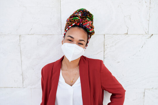 Young woman with protective face mask and headscarf staring while standing against white wall