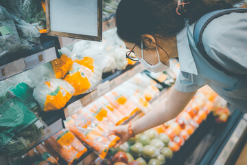Yong woman wears face mask shopping, selecting fruits, vegetables, ingredients or food stuff at grocery store or supermarket for cooking at home after working. New normal activity during covid-19.