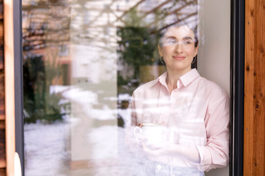 Smiling female entrepreneur with coffee mug looking away seen through glass window