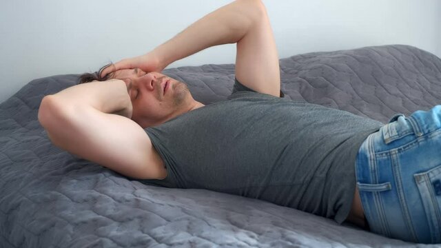 Young man is waking up in bed wearing in jeans and t-shirt after party. Man with morning hangover at home. He sleeps on the unmade bed. The life of a person with bad habits, alcoholism.