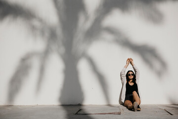 Young woman with arms raised sitting by palm tree shadow on white wall