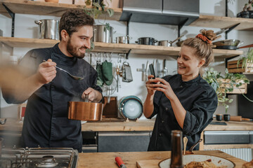 Young chef taking photo of colleague tasting soup while standing in kitchen