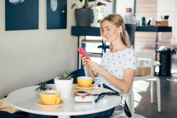 Portrait of a young beautiful caucasian stylish blonde sitting at a table in a coffee shop. Woman laughing while chatting in the mobile phone.