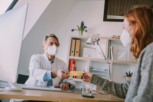 Male doctor wearing protective face mask giving vaccination certificate to patient while sitting at office