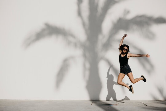 Young woman jumping with arms outstretched against shadow on white wall