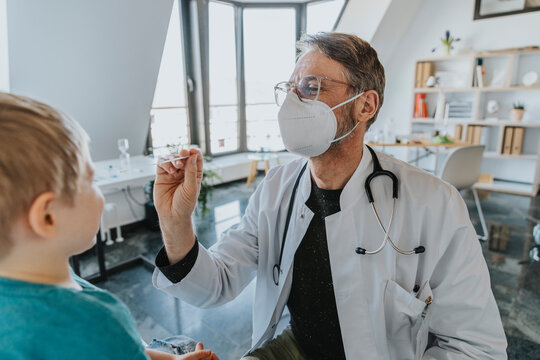 Male wearing protective face mask pediatrician taking boy nasal swab test while standing at clinic