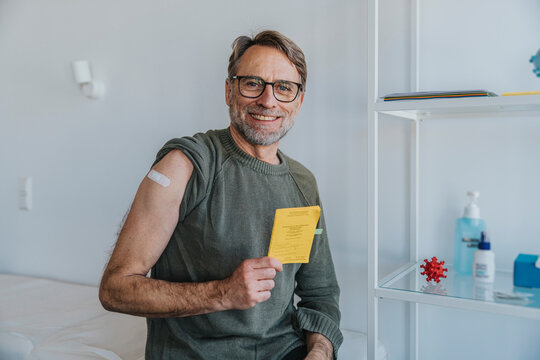 Smiling man with bandage on arm showing vaccination certificate while sitting at examination room