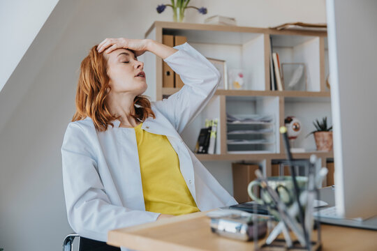 Frustrated Female Doctor Sitting With Head In Hand At Doctor's Office