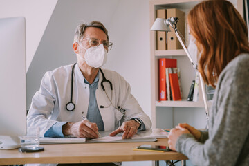Male general practitioner with face mask looking at patient while sitting at doctor's office
