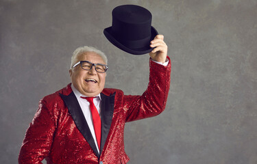 Happy mature man in glasses and fancy sequin jacket says hello and takes off black top hat. Funny elderly wedding toastmaster, conjurer, illusionist, announcer, showman or presenter greeting audience - obrazy, fototapety, plakaty