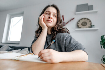 Thoughtful woman sitting at table while writing in notebook in living room