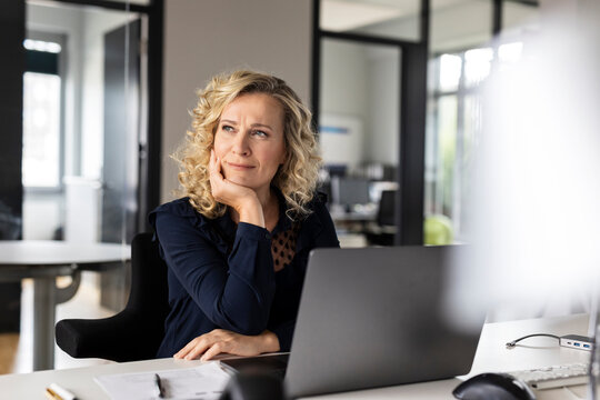 Thoughtful female business professional with head in hand looking away while sitting at desk in office