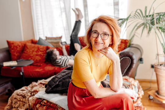 Smiling Woman Sitting On Couch At Home While Son Relaxing On Sofa In Background