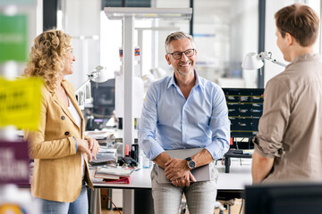 Smiling businessman with digital tablet discussing with colleagues while leaning at desk during meeting in office