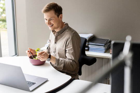 Smiling young businessman having lunch while looking at laptop in office
