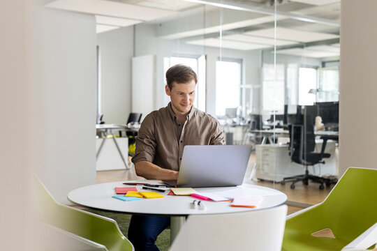 Smiling young businessman using laptop while sitting at table in open plan office