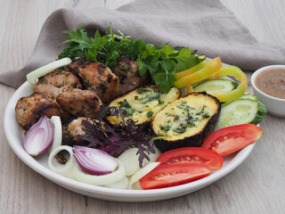 Shish kebab with herbs, fried stuffed potatoes in the skin, fresh various vegetables in a white plate on the table, closeup. Delicious rustic food with grilled meat barbecue and natural products
