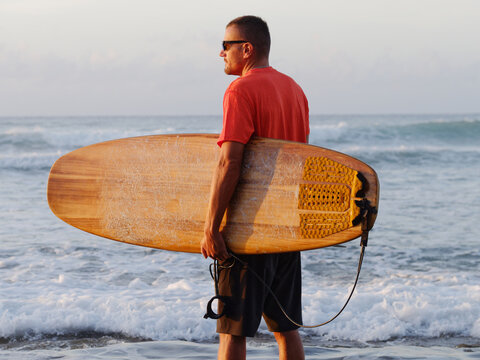 Man wearing orange t-shirt and black shorts, standing with a brown surfboard at the surf beach. Sunrise. 