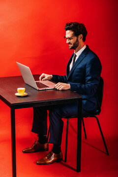 Smiling Businessman Using Laptop At Desk By Red Background