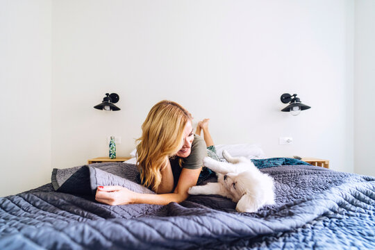 Blond woman playing with puppy on bed against white wall at home